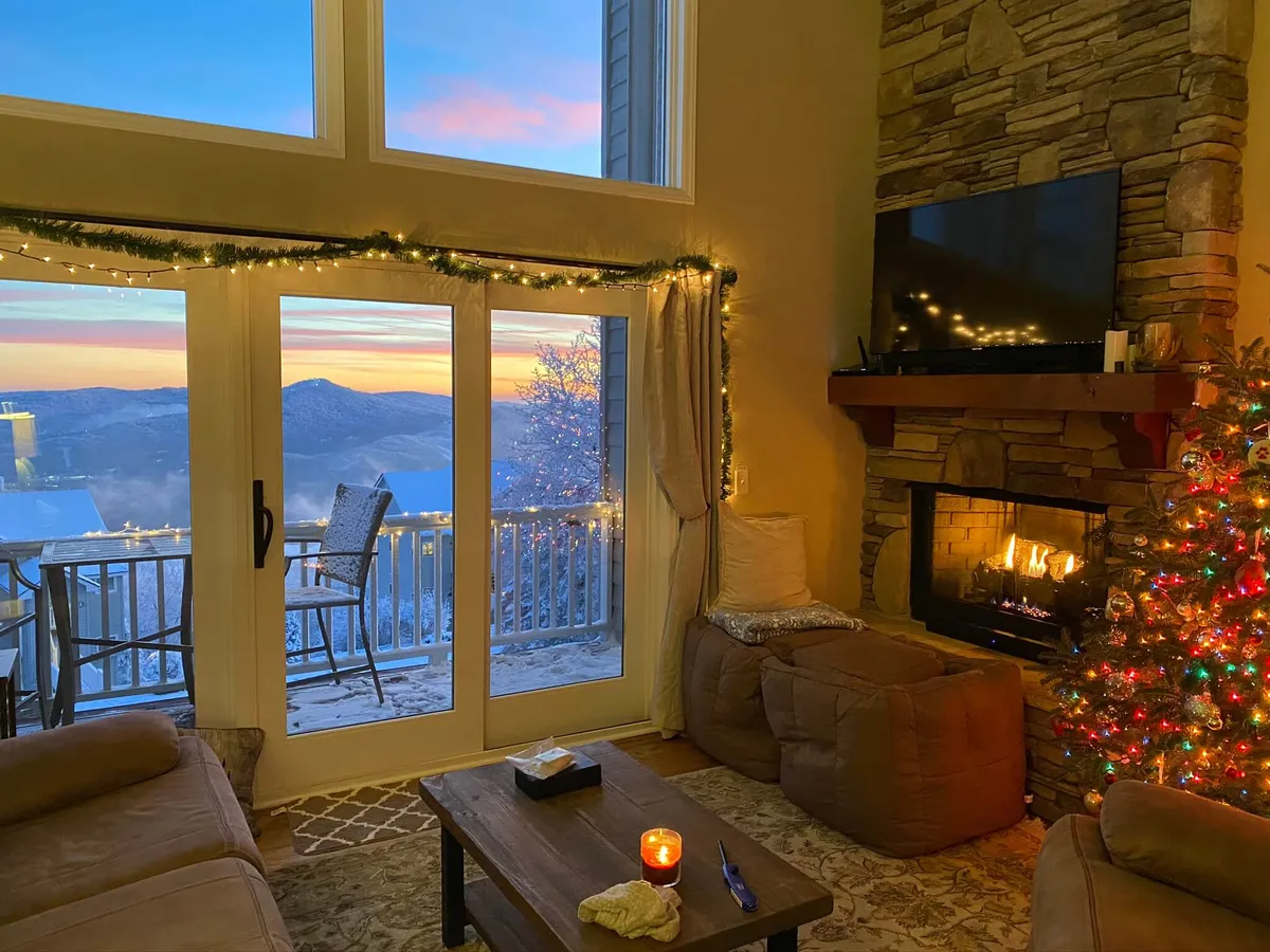 A warmly lit interior with a prominent fireplace, large windows letting in natural light, a decorated Christmas tree, and comfortable seating arranged for social gatherings. The inviting atmosphere suggests a space ideal for relaxing or entertaining guests, with clear sightlines that hint at a connection between living and dining areas.