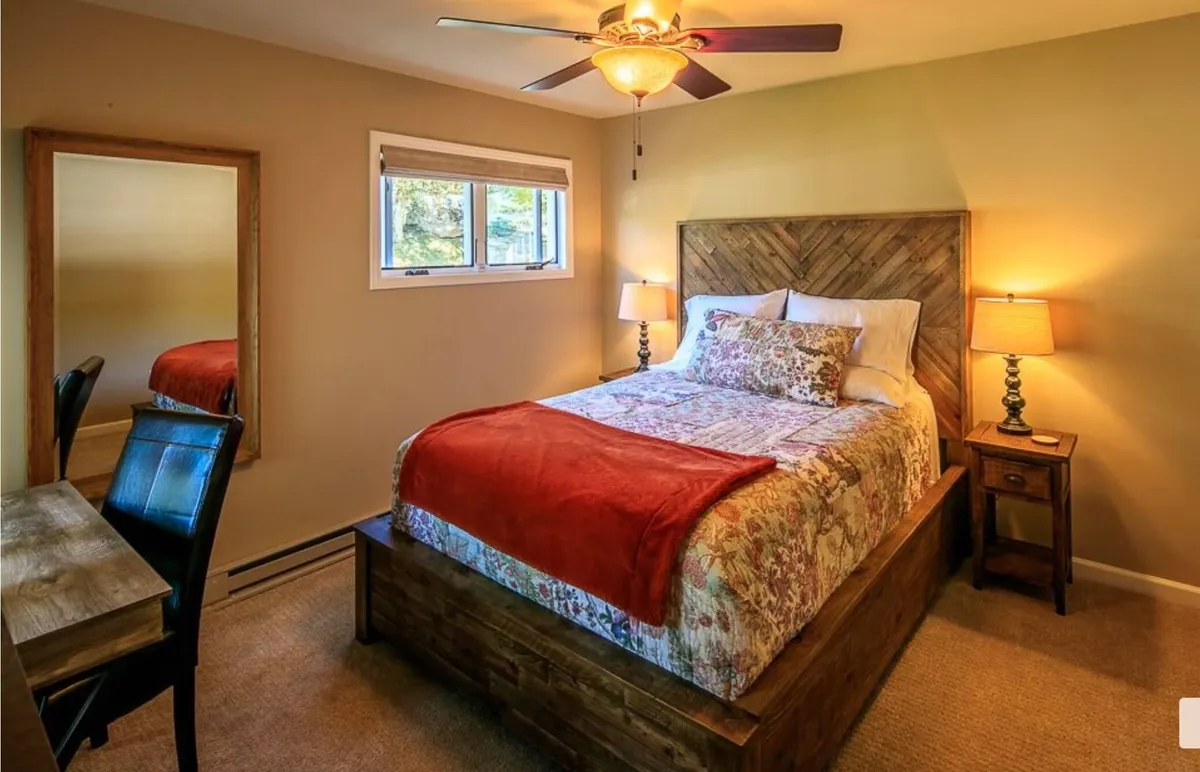 A warmly lit bedroom featuring a bed dressed in colorful, inviting bedding, flanked by matching nightstands each holding a lamp. A large mirror on one wall reflects the room's natural light, and a small desk offers a space for reading or working. The overall mood is cozy and welcoming, suggesting a relaxing, restful atmosphere.
