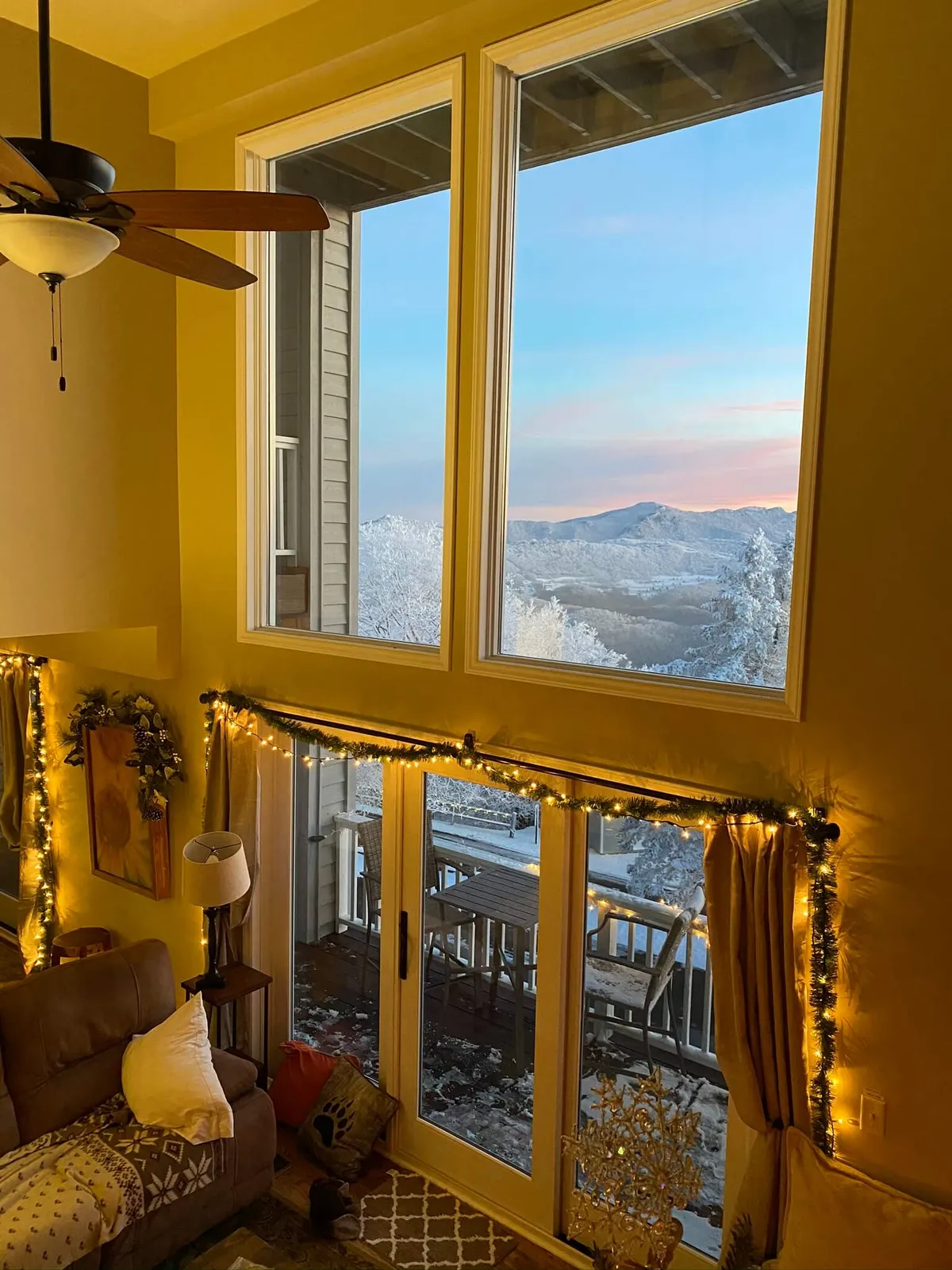 A cozy living area with a large window framing a stunning mountain view, a beautifully decorated Christmas tree, and a plush couch. Soft natural light pours in, highlighting the tranquil mountain landscape visible from inside. The decor evokes warmth and togetherness, making it an inviting spot for relaxation and socializing.