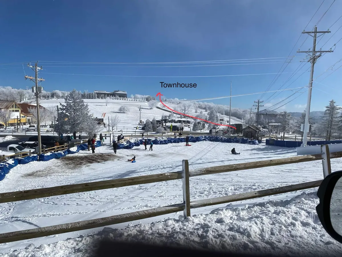 A lively exterior scene featuring a snow-covered hill adjacent to the townhouse. Families and friends are engaged in sledding and snowball fights, with the townhouse visible in the background. Blue tarps and surrounding trees add to the winter resort atmosphere, while groups enjoy building snowmen and sipping hot cocoa.