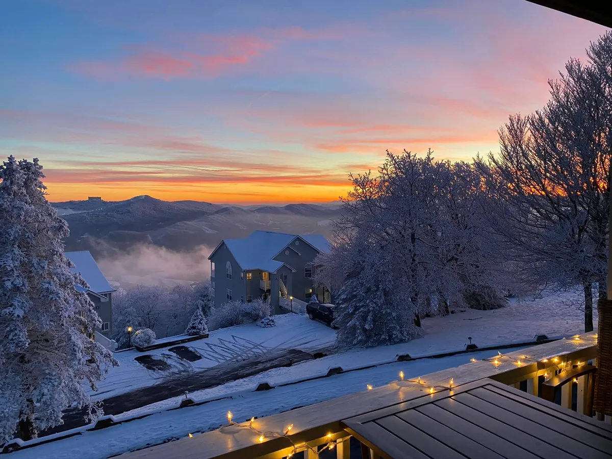 A scenic view from the deck looking out over snow-covered trees and distant mountains, with a colorful sunset sky. Warm lights on the deck suggest an inviting space for evening relaxation or al fresco dining, perfectly capturing the transition between inside and outside.