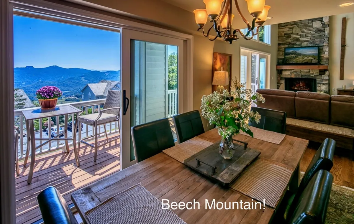 An elegant dining area featuring a wooden table set for a meal, with expansive windows revealing a panoramic view of the mountains. The warm glow of a nearby fireplace enhances the atmosphere, and the open layout hints at seamless flow between living, dining, and kitchen spaces.